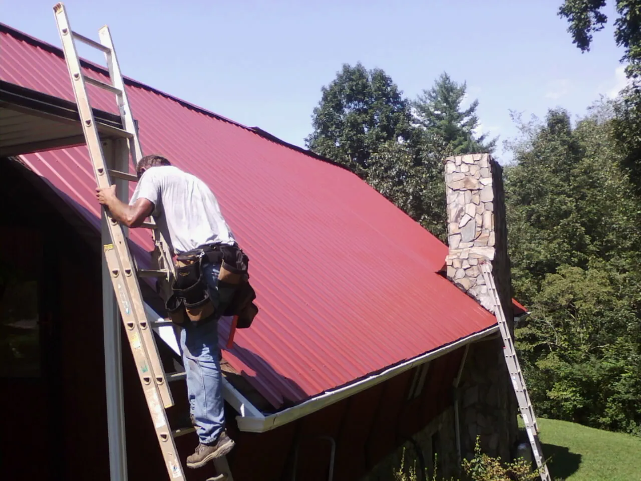 Roofer on a ladder beside a red metal roof and stone chimney — Soffit & Fascia Repair by Roofing Brothers in Sherman Oaks